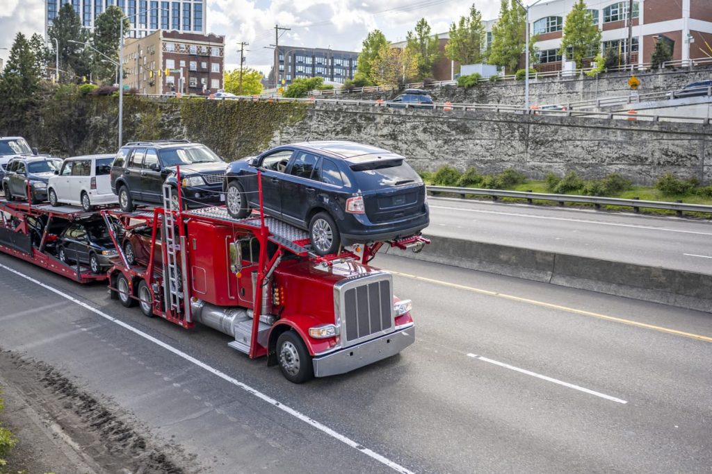 Red powerful big rig car hauler semi truck with loaded modular semi trailer driving on the highway road at the city limit going to point of destination