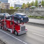 Red powerful big rig car hauler semi truck with loaded modular semi trailer driving on the highway road at the city limit going to point of destination