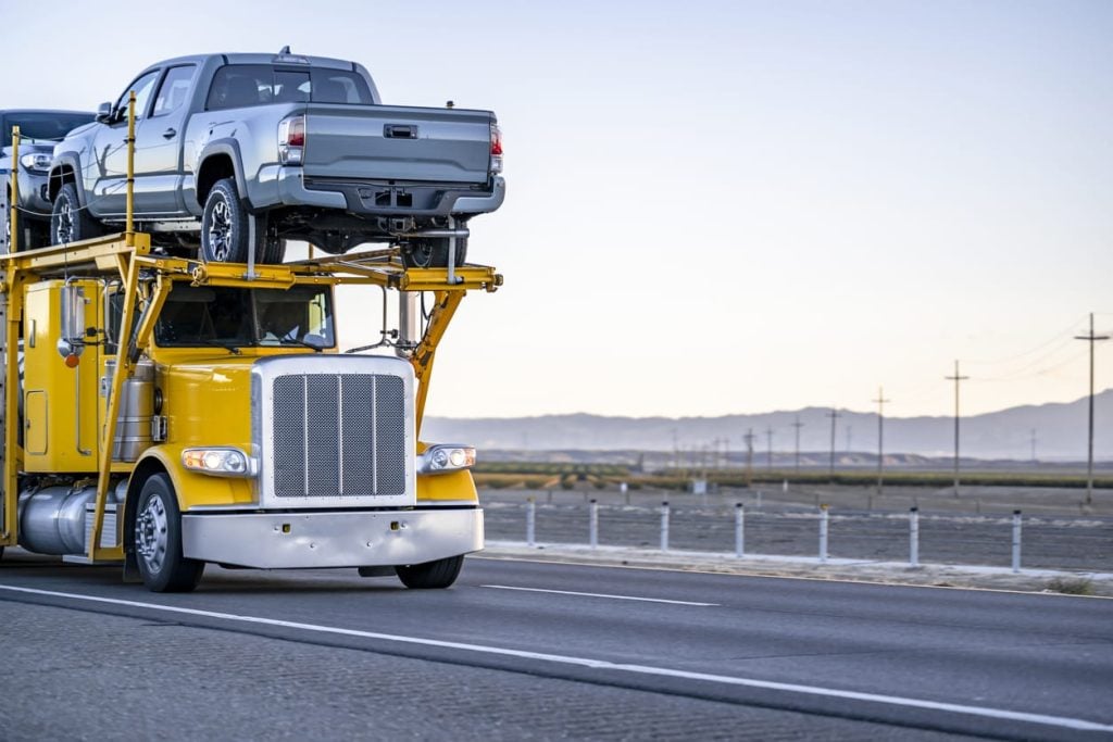 Bright yellow industrial standard big rig car hauler semi truck transporting pick up trucks on two level modular semi trailer driving on the road at twilight