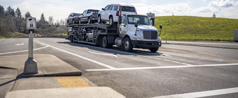 Compact white big rig car hauler semi truck transporting cars on the two level module semi trailer standing on the crossroad intersection with crosswalk