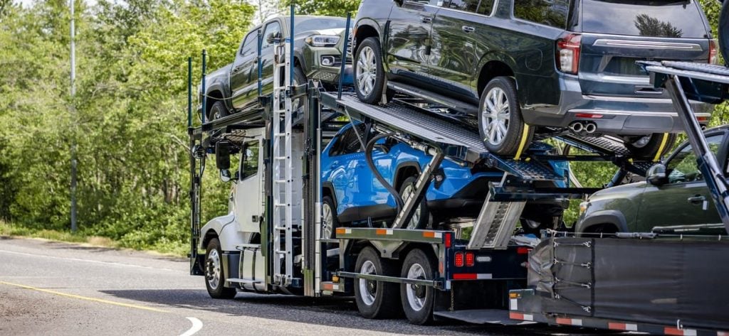 Powerful white big rig car hauler semi truck transporting cars on the two level modal semi trailer running on the road with trees on the side