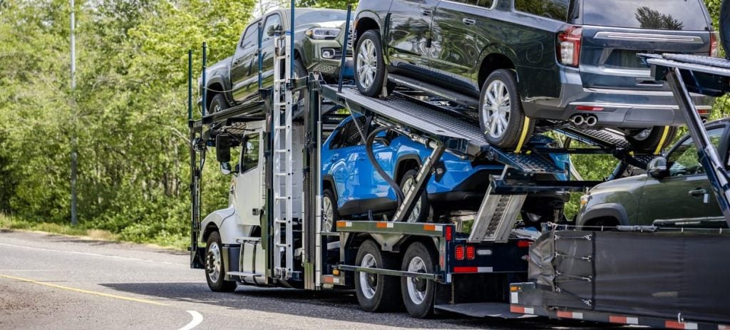 Powerful white big rig car hauler semi truck transporting cars on the two level modal semi trailer running on the road with trees on the side