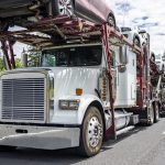 Car hauler white big rig classic semi truck transporting cars on the modular two level semi trailer running on the turning summer road