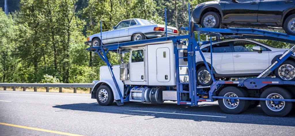 White classic car hauler big rig semi truck with blue modular semi trailer transporting cars running on the green highway road with forest and waterfall