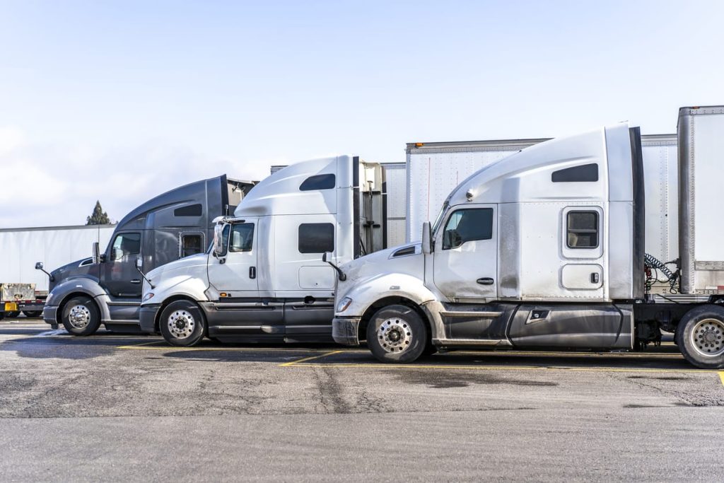 Profiles of big rigs semi trucks with semi trailers stand on wide parking lot of truck stop