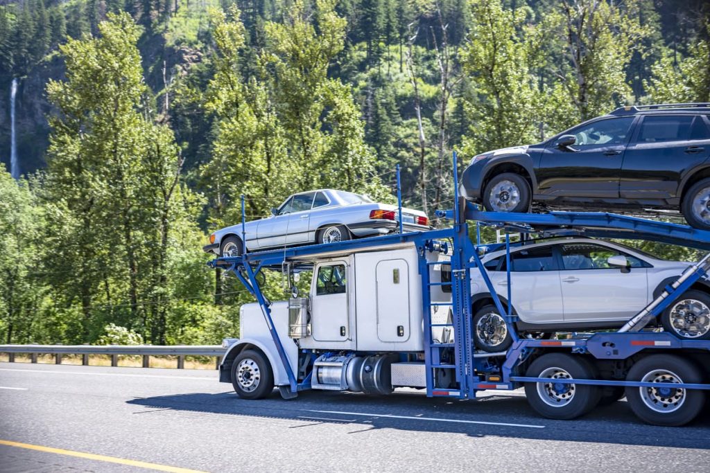 White classic car hauler big rig semi truck with blue modular semi trailer transporting cars running on the green highway road with forest and waterfall
