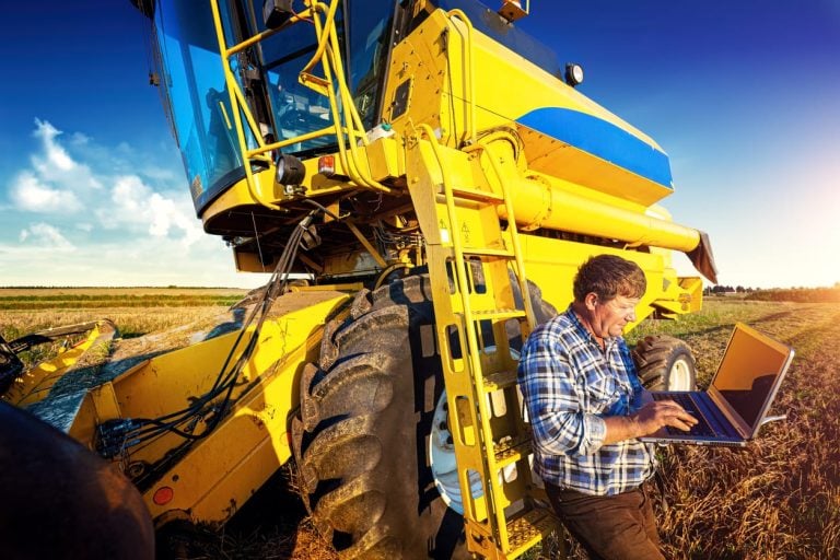 A farmer leaning against a combine harvester uses a computer during harvest.