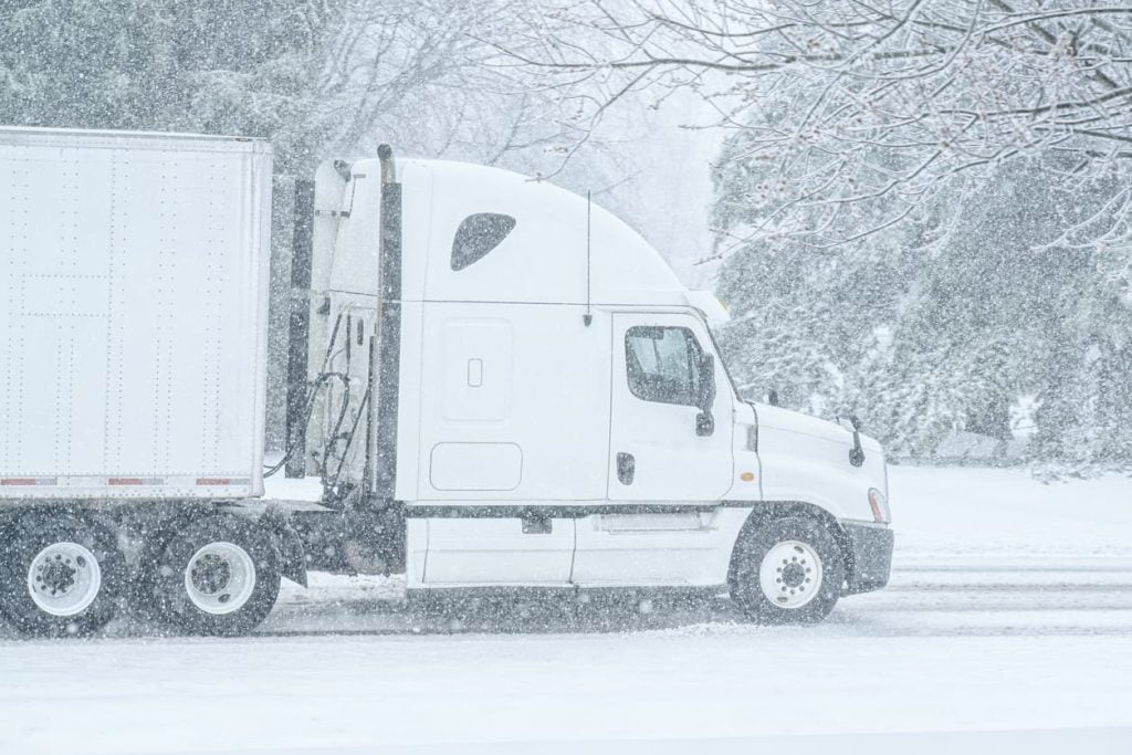 A white semi tractor trailer on a country road during a snow storm