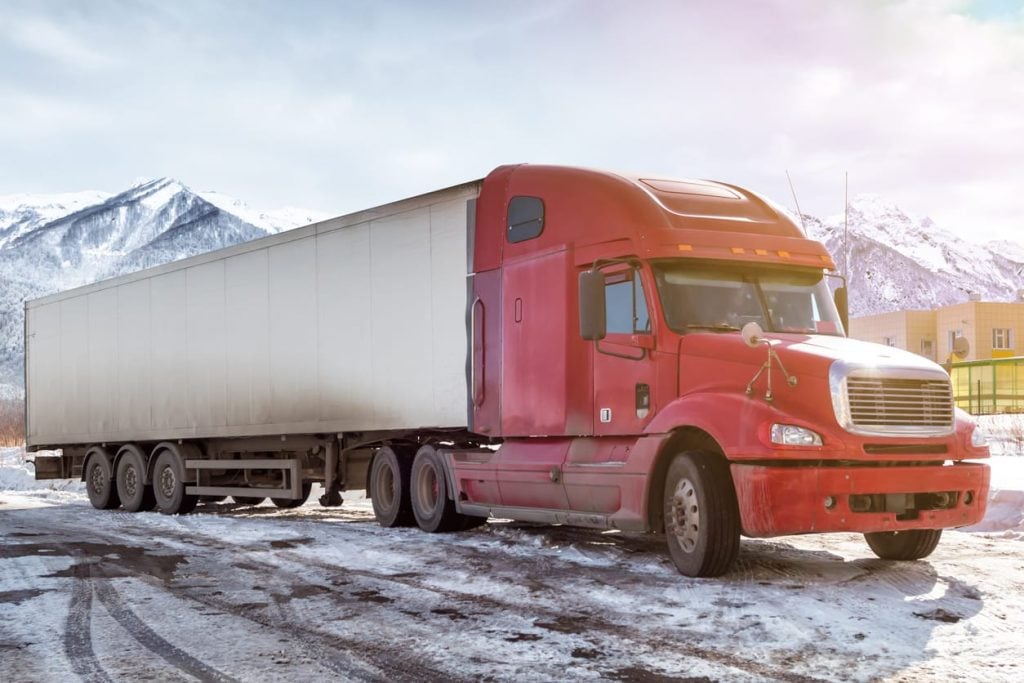 Red long-distance bonnet truck with a white semitrailer at winter on the background of high scenic mountains