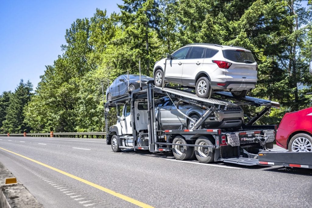 Compact powerful white big rig car hauler semi truck transporting cars on two hydraulic modular semi trailer running on the wide highway