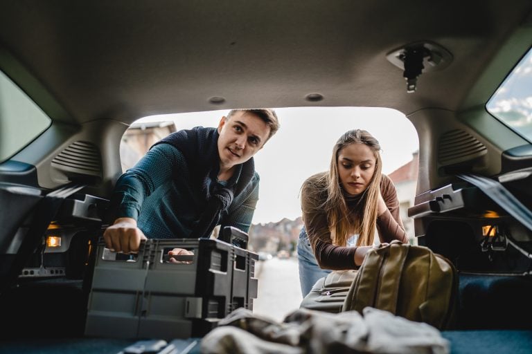 Two people couple students travel concept man and woman taking luggage baggage suitcase and other stuff and belongings from the back of car while moving into dormitory on college campus real people