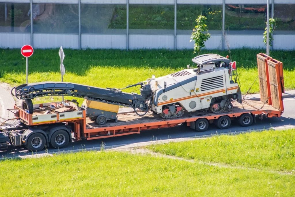 Transportation of equipment for cutting and removing old asphalt pavement for road repair on a truck platform of a truck trailer on a highway in a city, side aerial view.