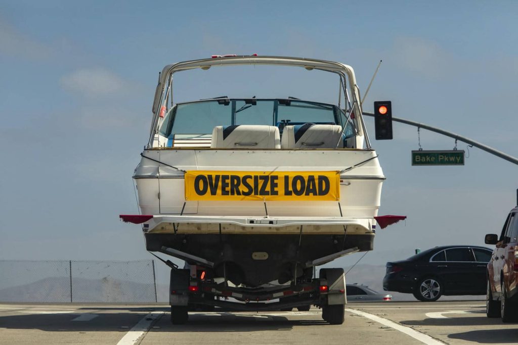 A large white motor boat being hauled on a trailer with a yellow sign stating Oversize Load