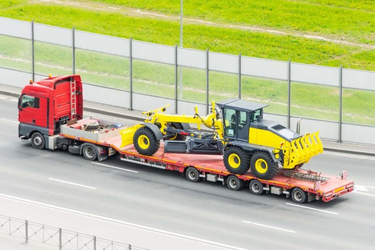 Grader tractor on transportation trailer truck with long trailer platform on the highway in the city.