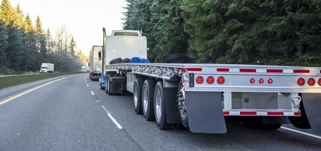 Big rig long haul semi truck with empty flat bed semi trailer running on the winter road in convoy with another semi trucks