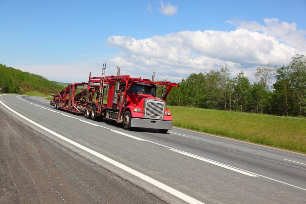 Red car hauler semi truck with empty trailer on a highway between green hills