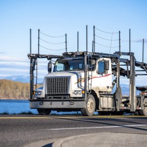 White car hauler semi truck driving a narrow road along a river with hills in the distance