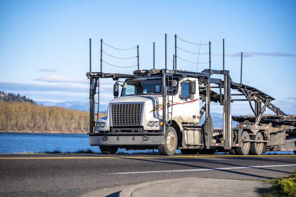White car hauler semi truck driving a narrow road along a river with hills in the distance