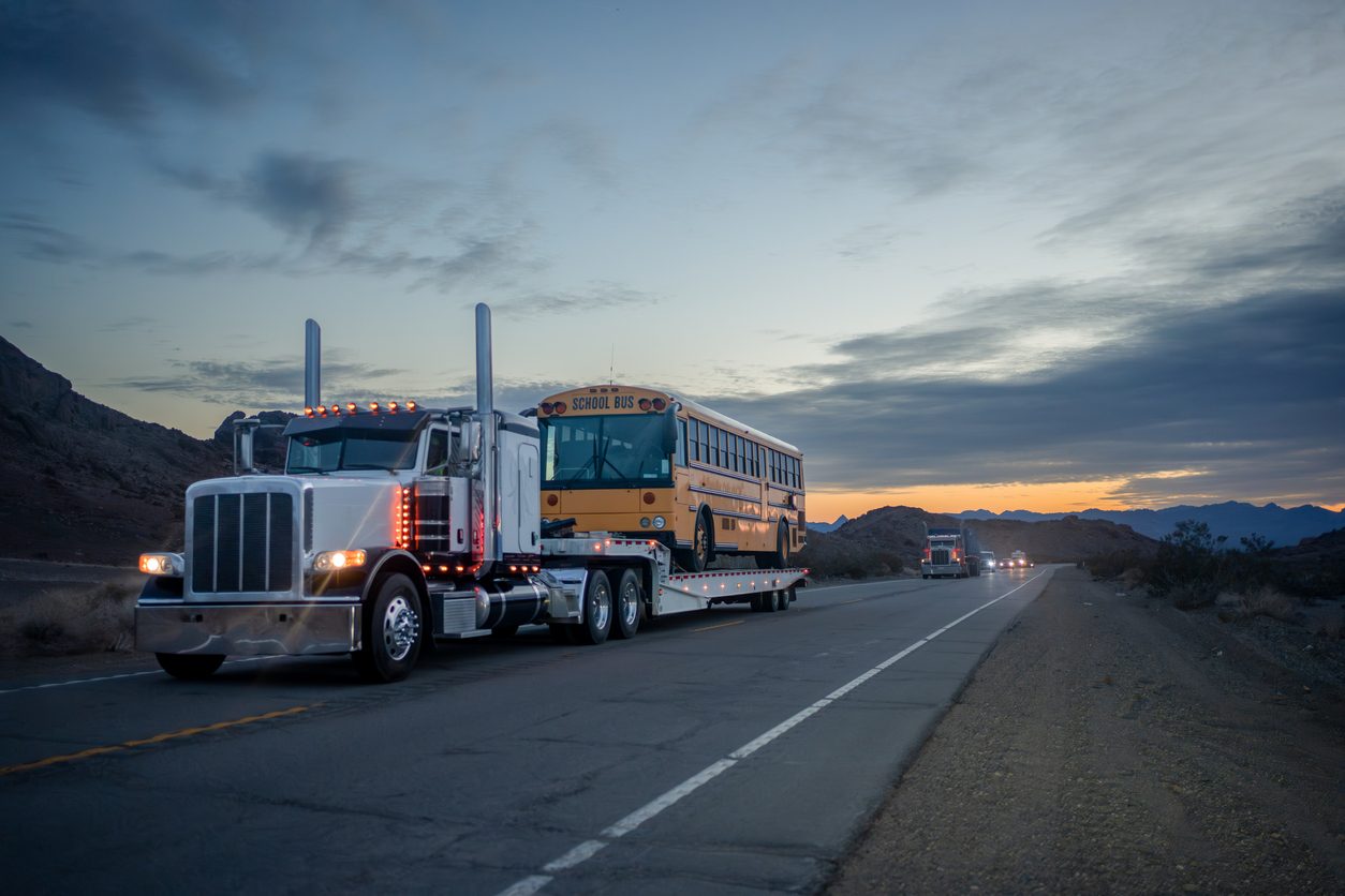 Flatbed truck hauling a school bus through the Arizona desert at dusk