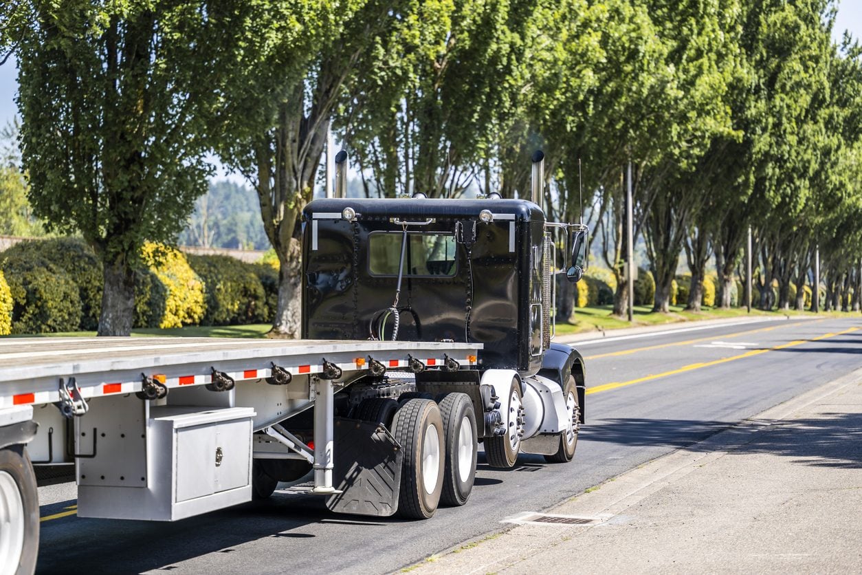 Black conventional tractor with a flatbed trailer on a tree lined street