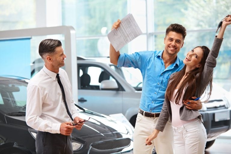 Couple with paperwork and keys beside a car in a dealership showroom