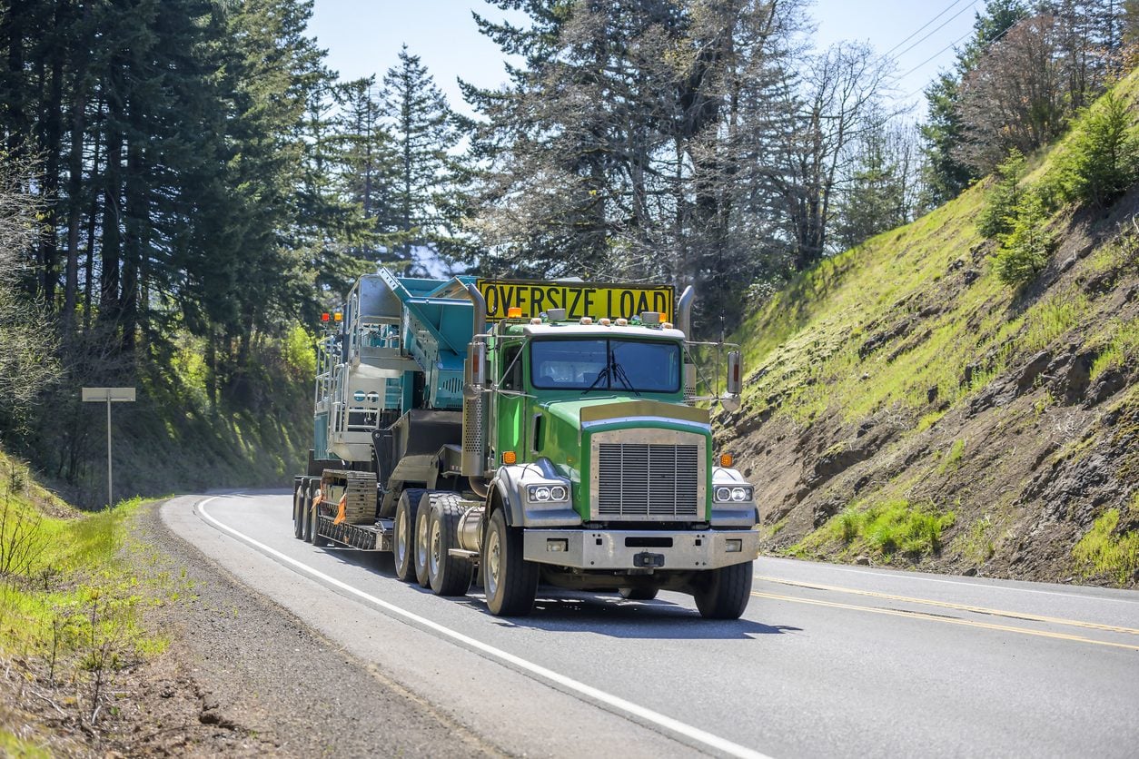 Step deck trailer carrying a crawler machine up a forested mountain highway
