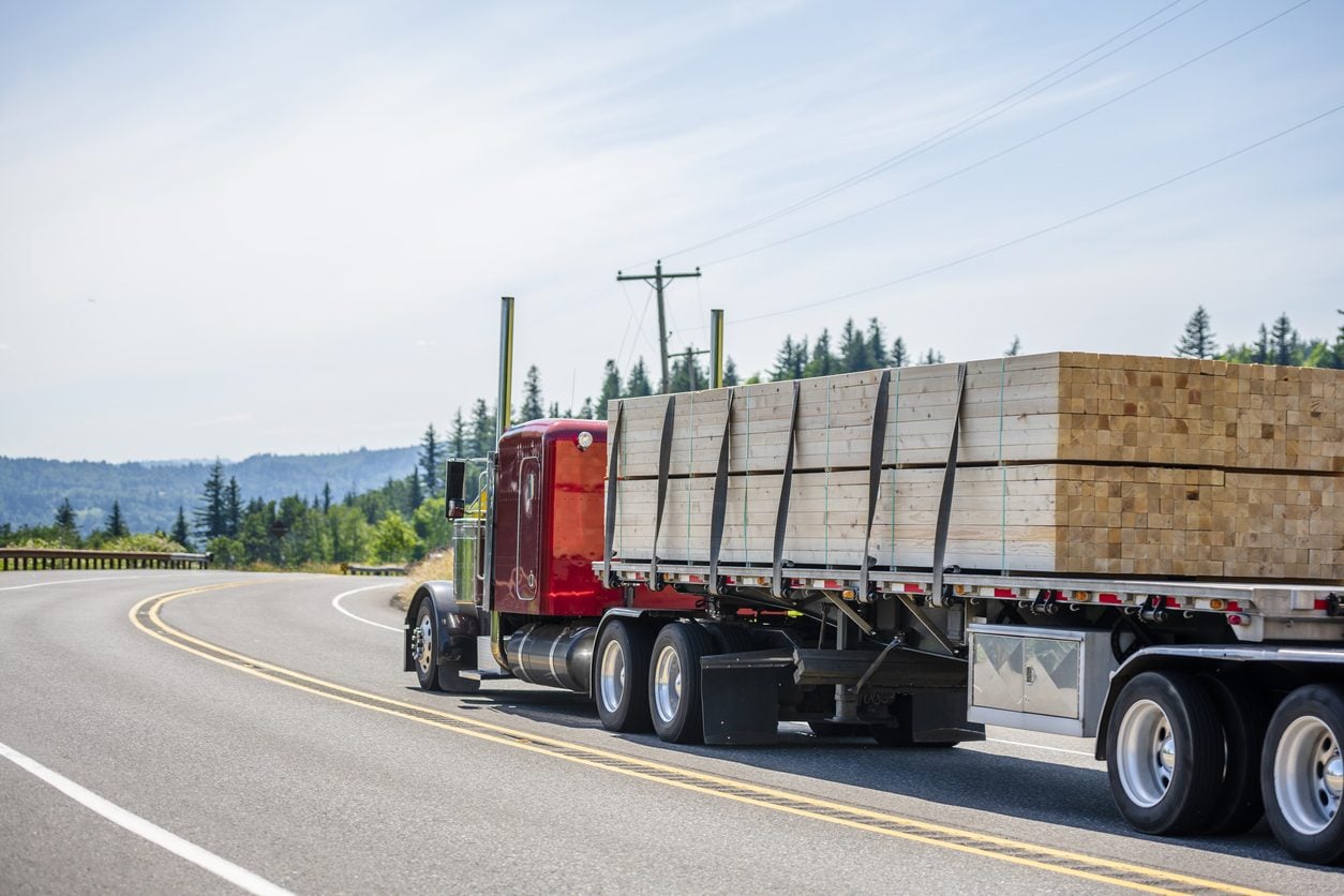 Red conventional tractor hauling lumber on a flatbed trailer