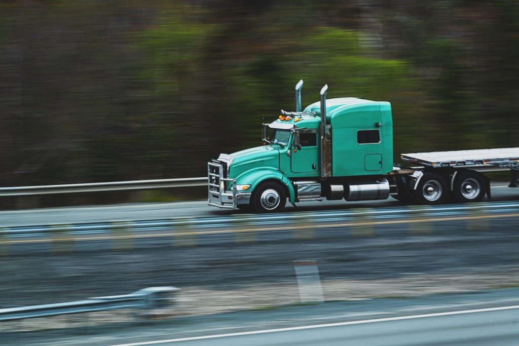 Teal conventional tractor with a flatbed trailer moving along a highway