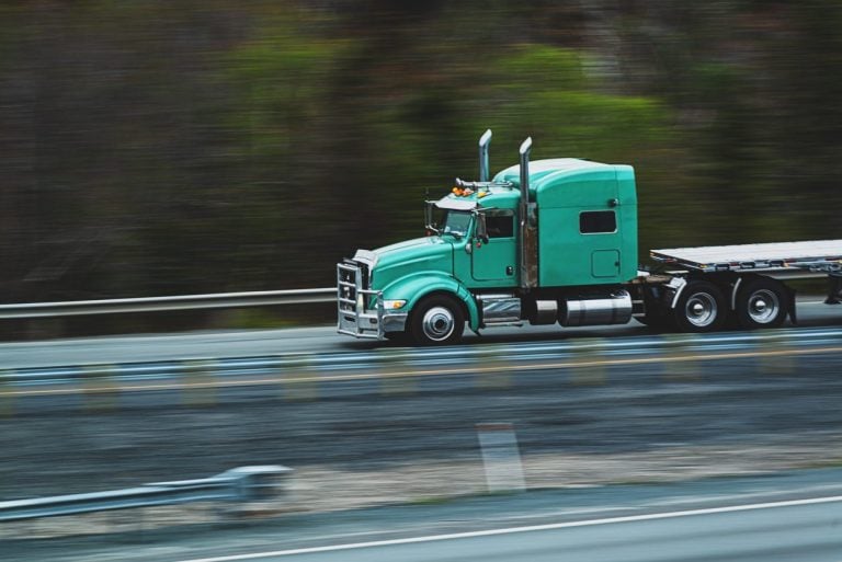 Teal conventional tractor with a flatbed trailer moving along a highway