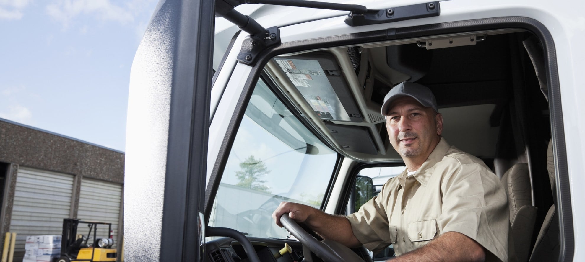 Commercial driver seated in the cab of a white truck with his hand on the wheel, ready to drive the unit to its destination