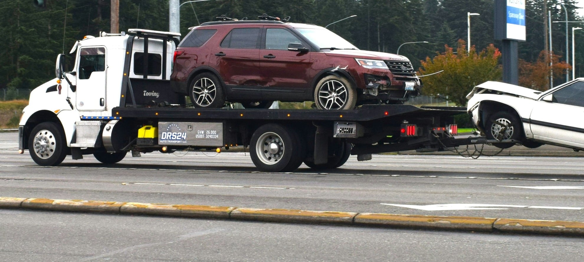 Dark red SUV secured on the deck of a white flatbed carrier with a second non running sedan hitched behind on a US road