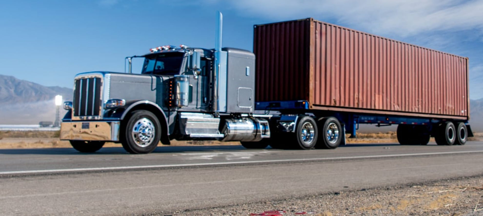 American semi truck hauling a rust red forty foot shipping container on a chassis along an open highway
